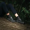 Gros plan sur les jambes d’un modèle portant les Mocassins Roma en cuir noir de Poema Paris, assise sur un tronc d’arbre, chaussures femme éco-responsables.