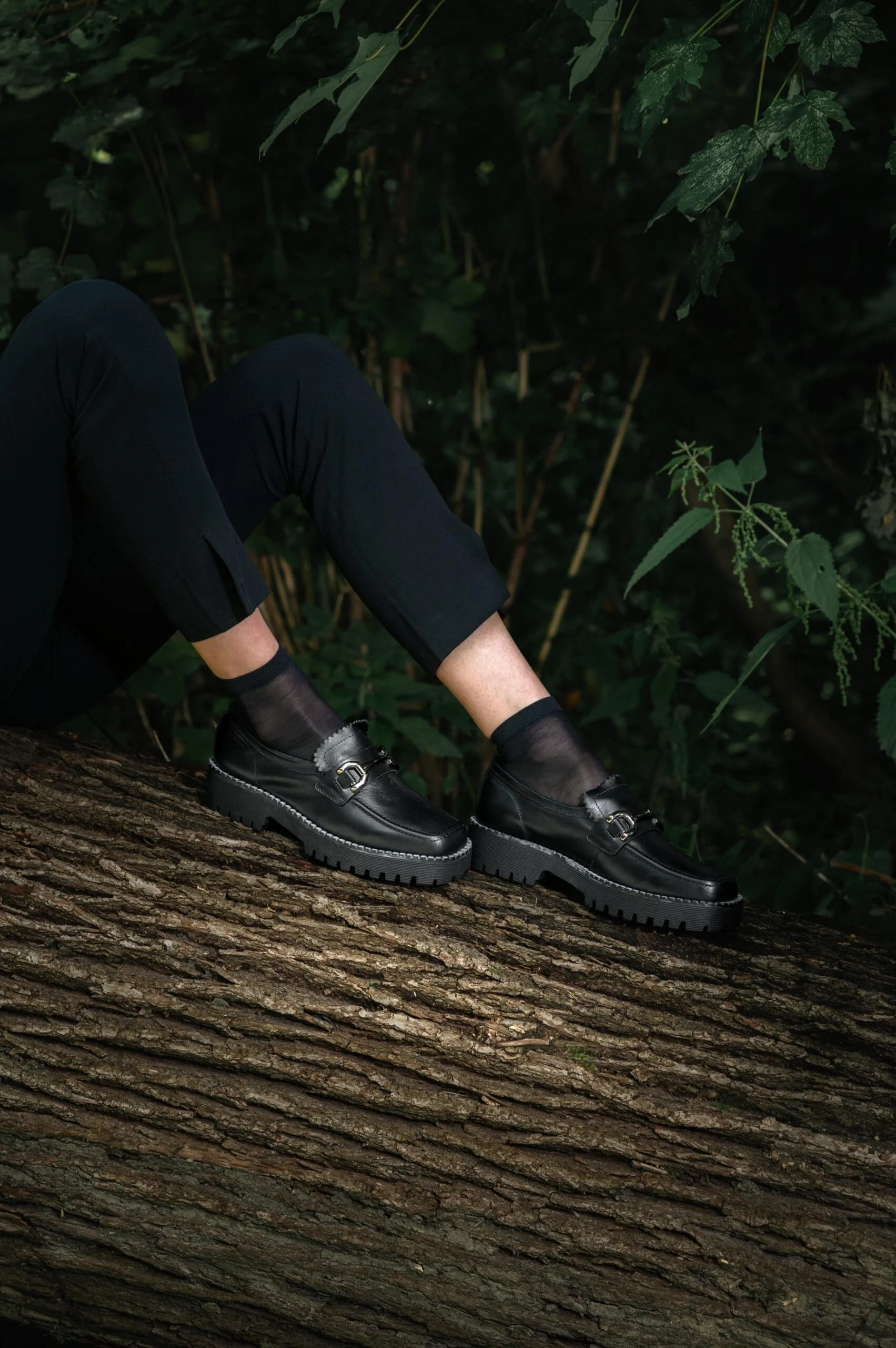 Mocassins en cuir noir éco-responsables de Poema Paris, portés par une modèle assise dans une forêt, chaussures femme élégantes et durables.